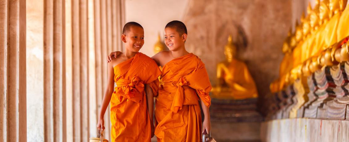 Two young Buddhist monks in traditional orange robes walking side by side in a temple corridor.
