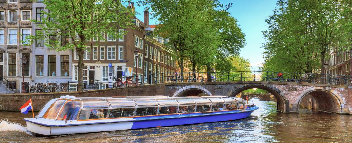 Glass-covered canal boat passing beneath a brick bridge along a tree-lined canal in Amsterdam