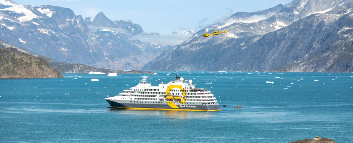 Ultramarine expedition ship navigating icy Arctic waters surrounded by rugged mountains and floating ice, with helicopters overhead and zodiacs beside the vessel.
